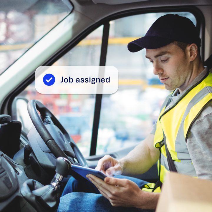 A delivery driver sitting in the drier seat of a van wearing a high vis jacket. He is looking at a mobile device while the van is stationary. Overlayed on the image is a notification saying 'Job assigned'